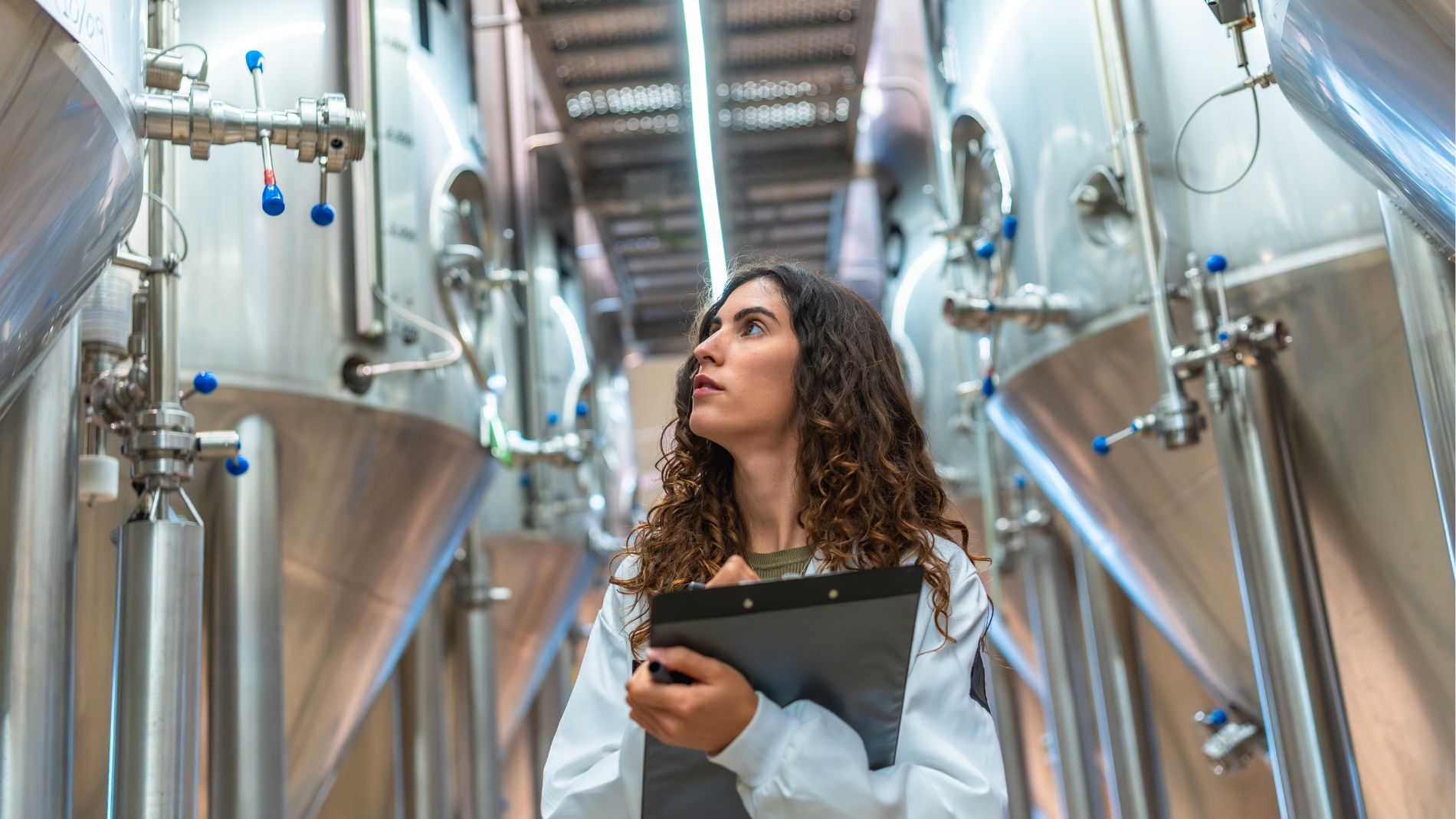Frau im weißen Laborkittel inspiziert Fermentationstanks in einer modernen Lebensmittelproduktionsanlage.