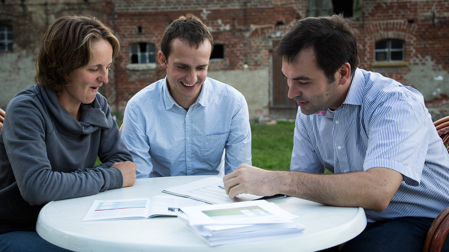Three people talking at table in front of old farmhouse – discussing agricultural topics