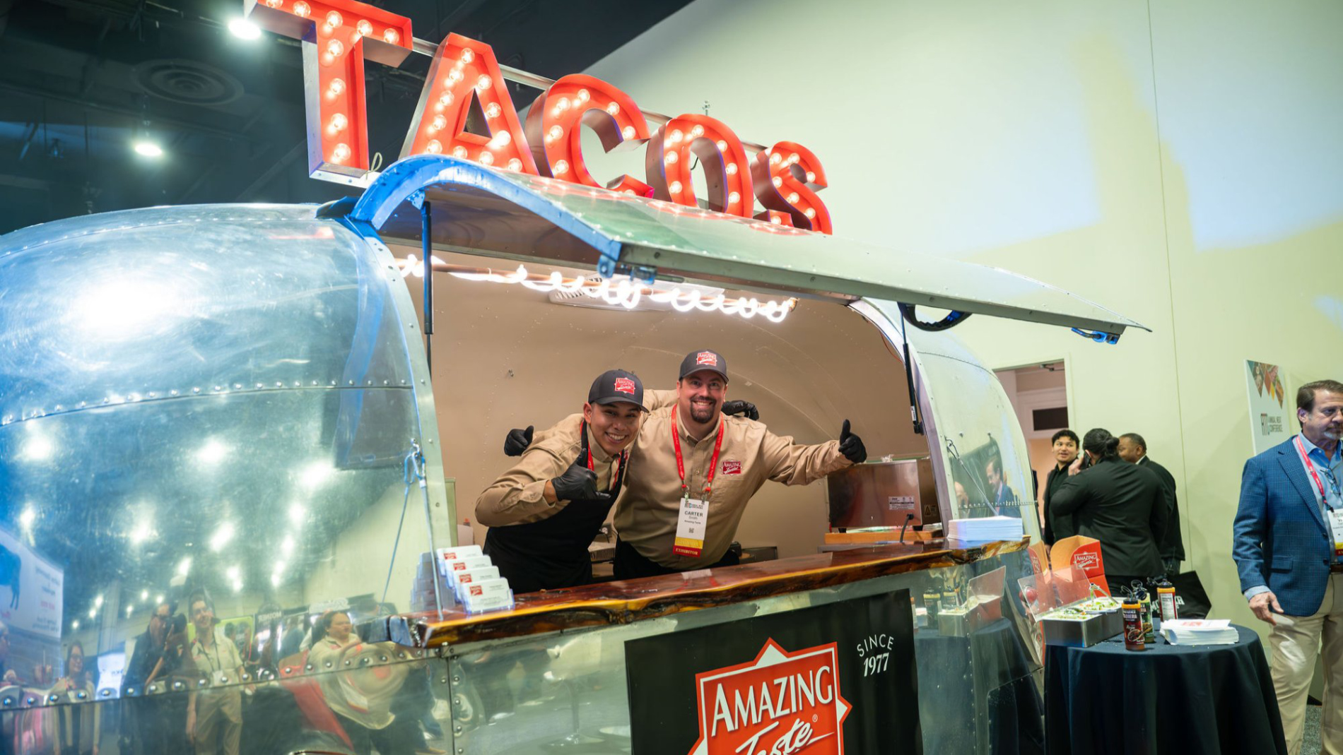 Zwei Standmitarbeiter begrüßen Besucher aus einem Food-Trailer mit leuchtendem „Tacos“-Schild auf einer Messe.