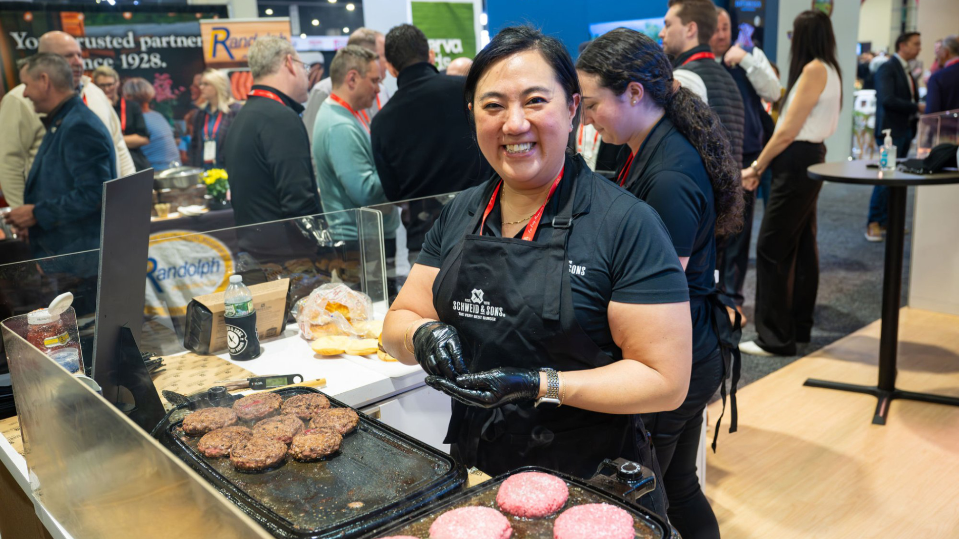 Eine Standmitarbeiterin bereitet auf einer Messe frische Burger zu, während sich im Hintergrund zahlreiche Besucher an den Ständen austauschen.